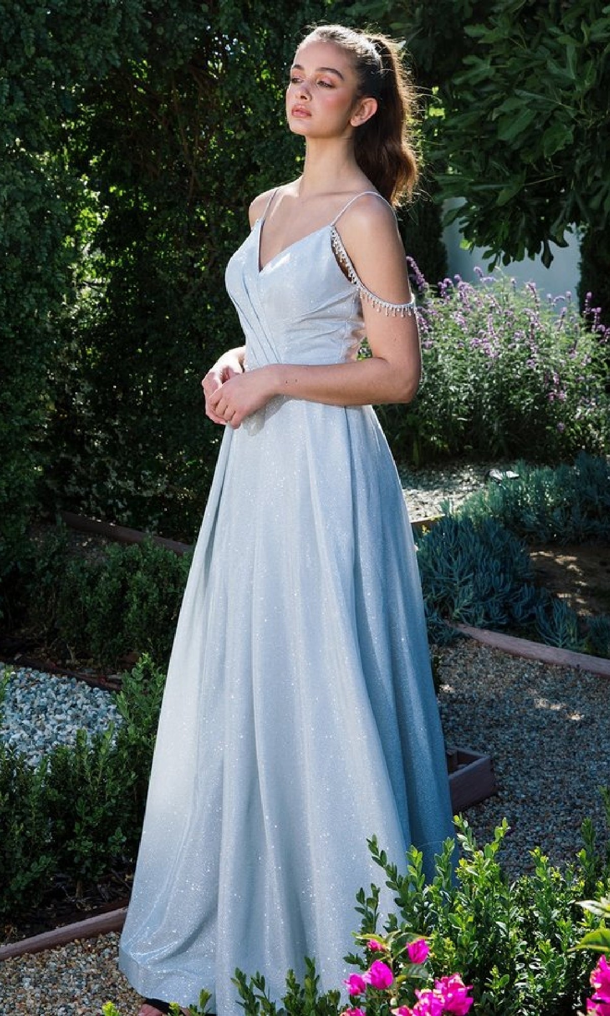 A woman stands in a garden wearing the Eureka 9800 Lace-Up Long Shimmer Prom Dress, light blue and sleeveless, with thin straps and decorative beading on one shoulder.